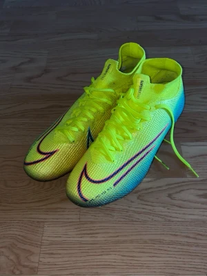 Nike Mercurial Superfly Elite fotbollsskor - Säljer ett par Nike Mercurial Superfly fotbollsskor i neon-gult med blå och lila detaljer. Skorna har högt skaft, snörning och är tillverkade i ett lätt, syntetiskt material för bästa grepp och känsla på planen. Medföljer matchande gympåse.