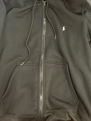 Svart hoodie från Polo Ralph Lauren - Snygg svart hoodie med dragkedja från Polo Ralph Lauren. Klassisk design med vit broderad logga på bröstet, två fickor framtill och justerbar huva med snören. Tillverkad i mjukt bomullsmaterial som är skönt att bära.