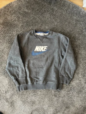 Grå sweatshirt från Nike - Klassisk grå sweatshirt från Nike med stor vit och blå logga på bröstet. Tröjan har rund halsringning, ribbade muddar vid ärmslut och nederkant samt en relaxed passform. Perfekt för en sportig och avslappnad stil. Storlek 146 12 år.