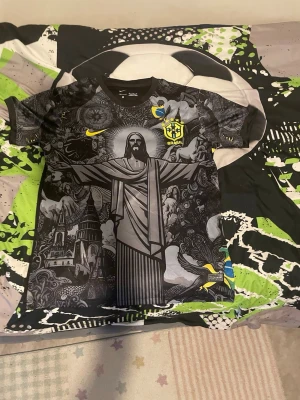 Svart Brasilien fotbollströja Nike - Unik svart fotbollströja från Nike med Brasilien-emblem och gult Nike-märke. Framsidan har ett stort tryck av Kristusstatyn och detaljerade illustrationer i grått och svart. Korta ärmar med brasilianska flaggan och grafiska mönster. Perfekt för dig som älskar fotboll och streetwear.