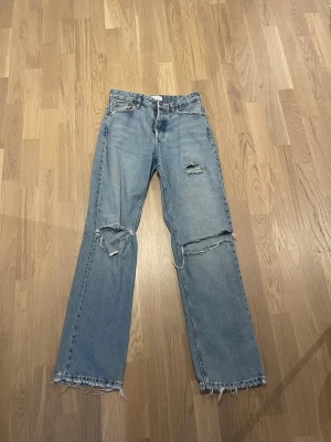 Ljusblå raka jeans med slitningar - Säljer ett par ljusblå raka jeans med slitna detaljer och hål på knäna. Jeansen har fransiga benslut och klassisk femficksmodell. Perfekta för en avslappnad och trendig look. .