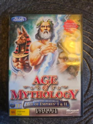 Age of Mythology PC-spel - Age of Mythology är ett strategispel för PC där du styr mytologiska civilisationer och gudar. Omslaget har färgstarka illustrationer av gudar och mytiska varelser. Spelet kommer i ett klassiskt plastfodral och är utvecklat av Ensemble Studios.