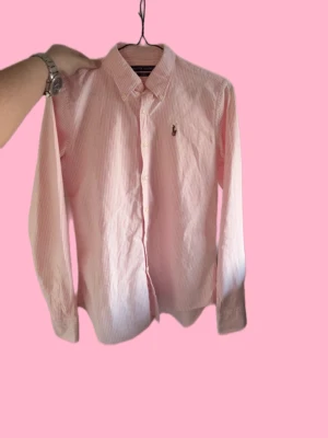 Rosa randig skjorta från Ralph Lauren - Säljer en slim fit skjorta från Ralph Lauren i rosa och vitt med tunna ränder. Skjortan har klassisk krage, knäppning framtill och den ikoniska broderade loggan på bröstet. Perfekt för dig som gillar preppy stil och snygga detaljer. Jag har aldrig använt den jag köpte den helt ny men den är liten på mig så det därför jag säljer den för så billigt. Tveka inte att fråga eller ge pris bud är flexibel med det 