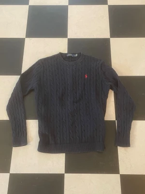 Mörkblå kabelstickad tröja Polo Ralph Lauren - Kabelstickad mörkblå tröja från Polo Ralph Lauren med rund halsringning och röd broderad logga på bröstet. Tröjan har långa ärmar och ribbade muddar vid ärmslut och nederkant. Klassisk och tidlös design som passar till mycket. QR koden är härmed lite sliten, men annars perfekt 👌 Skriv vid funderingar! 💫 Pris kan diskuteras 🏆 