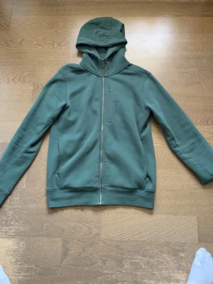 Grön hoodie från Sail Racing i mycket gott skick - Mörkgrön hoodie från Sail Racing med dragkedja framtill och broderad logga på bröstet. Hoodien har en mjuk fleeceinsida, huva och två sidofickor samt en bröstficka med dragkedja. Perfekt för en avslappnad och sportig stil. Tröjan kosta från 1600 och fick i present men har inte använt den så säljer nu för ett riktigt bra pris.