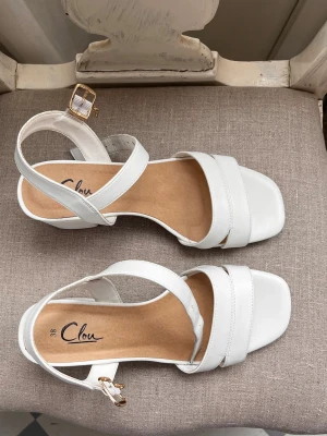 Vita sandaler med blockklack från Clou -  vita sandaler från Clou med breda remmar och fyrkantig tå. En stabil blockklack och justerbart spänne i guld. Använt en gång 