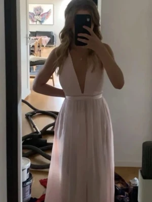 Ljusrosa långklänning djup v-ringning - Elegant ljusrosa långklänning med djup v-ringning och markerad midja. Klänningen är ärmlös och har ett flytande, lätt tyg som ger en romantisk känsla. Perfekt för bal eller student. Materialet känns som polyester eller liknande.