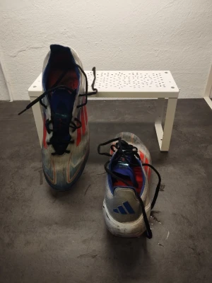 Adidas fotbollsskor blå/röd - Säljer ett par Adidas fotbollsskor med snygg design i blått, vitt och röda detaljer. Skorna har svarta snören, låg profil och mönstrad sula med dobbar för bra grepp på planen. Perfekta för dig som vill sticka ut på fotbollsplanen.