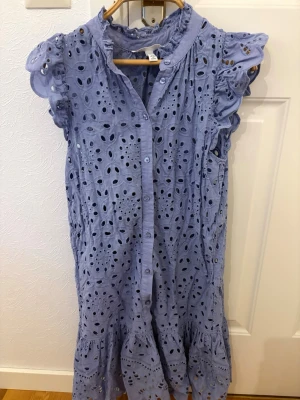 Klänning - Söt blå klänning i bomull med broderade hålmönster och volangdetaljer. Kort ärm med volang, knappar framtill och lätt rynkad nederdel. Perfekt för sommarens varma dagar