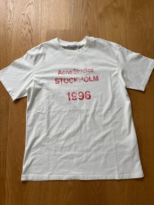 Vit Acne Studios t-shirt med tryck - Vit t-shirt från Acne Studios med rött tryck på bröstet där det står 'Acne Studios STOCKHOLM 1996'. Klassisk rund hals och korta ärmar. Tillverkad i mjuk bomull för en skön och avslappnad känsla.