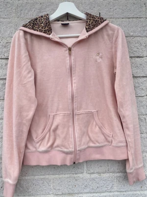 Rosa velour zipup- hoodie med leopardmönster - Säljer en ljusrosa hoodie i mjuk velour från Ginatricot. Huvan har leopardmönstrat foder och dragkedja framtill. Två fickor fram och broderad rosett logga med guldfärgad detalj på bröstet. Den är från ginas äldre sortiment och finns därför inte att få ta på längre.