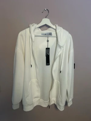 Vit hoodie med dragkedja från Stone Island - Säljer en fräsch vit hoodie från Stone Island med dragkedja framtill, huva med dragsko och klassisk logotyp-patch på ärmen. Hoodien har kängurufickor och är tillverkad i mjuk bomull för en skön känsla. Perfekt för en clean och stilren look.