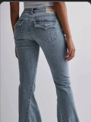 True Religion ljusblå bootcut jeans - Säljer ett par ljusblå bootcut jeans från True Religion med ikoniska bakfickor och dekorativa sömmar. Jeansen har låg midja, knappdetaljer på fickorna och är tillverkade i klassisk denim. Perfekta för en trendig och avslappnad look. Endast lite slitet ner till då dem är lite långa på mig! Men inte alls mycket, skriv för bild på de 💖