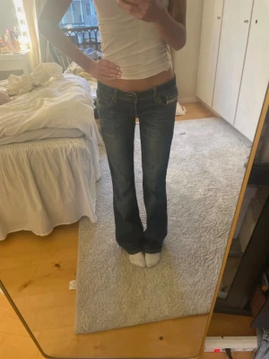 Blå bootcut jeans från Diesel - Säljer ett par klassiska blå bootcut jeans från Diesel med snygga slitningar och kontrastsömmar. Jeansen har låg midja, fem fickor och detaljerade nitar. Perfekta för dig som gillar en tidlös och avslappnad stil.