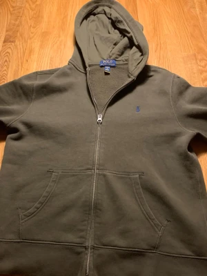 Olivgrön hoodie från Polo Ralph Lauren - Snygg olivgrön hoodie från Polo Ralph Lauren med dragkedja framtill och klassisk blå logga på bröstet. Hoodien har huva med dragsko och en stor känguruficka. Tillverkad i mjukt bomullsmaterial som är skönt att bära.