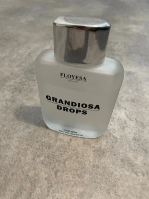 Grandiosa Drops parfym FLOVESA - En stilren parfymflaska från FLOVESA deluxe med namnet Grandiosa Drops. Flaskan är frostad vit med en stor, silverfärgad kork och tydlig svart text. Passar dig som vill ha en modern och maskulin doft i badrumshyllan.
