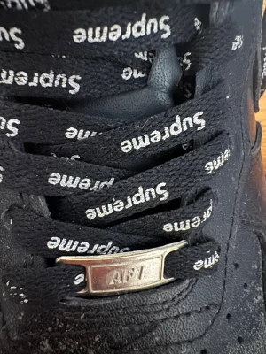 Nike Air Force 1 x Supreme svarta - Svarta Nike Air Force 1 Low i samarbete med Supreme. Skorna har svarta läderpaneler, svarta skosnören med Supreme-logga i vitt och en liten röd Supreme-tag på hälen. Klassisk Air Force 1-sula och metallbricka med 'AF1' på snörena. Perfekta för streetwear.