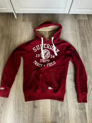 Vinröd Superdry hoodie  - Mysig vinröd hoodie från Superdry med vitt tryck framtill och stora fickor. Huva med vitt teddyfoder och breda vita dragsnören. Klassisk känguruficka och ribbade muddar. Hör av er för frågor o funderingar 
