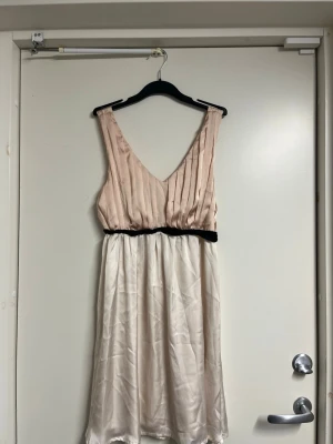 Ljus champagnefärgad omlottklänning - Elegant ljus champagnefärgad klänning med omlottdesign och djup v-ringning. Klänningen har ett svart band i midjan och är tillverkad i ett glansigt, mjukt material som ger en lyxig känsla. Perfekt för dig som vill ha en stilren och feminin look. (Jag har vanligtvis str xxs/xs och denna är något mellan S och M där av lite udda passform på mig) 