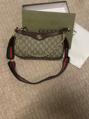 Gucci GG Supreme handväska med kedja - Snygg handväska från Gucci i beige GG Supreme canvas med bruna läderdetaljer och guldfärgad kedja. Väskan har en bred axelrem i grönt och rött, klassiskt Gucci-mönster och GG-logga i metall framtill. Perfekt accessoar för dig som gillar lyxig och trendig stil.