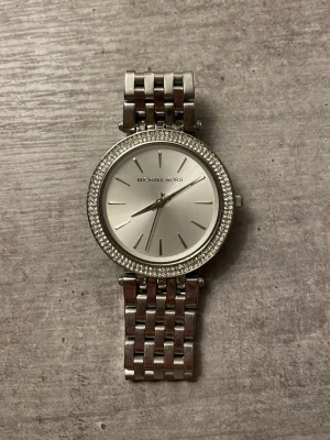 Silverfärgad klocka Michael Kors - Stilren klocka från Michael Kors i silverfärg med länkarmband och gnistrande stenar runt urtavlan. Klassisk rund form och minimalistisk design med tydliga markeringar på urtavlan. Perfekt accessoar för att lyfta din outfit.