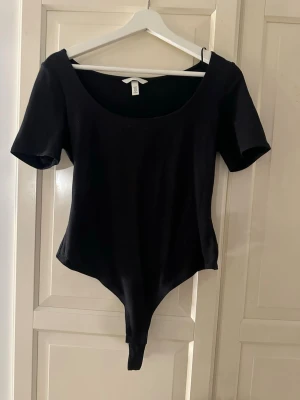 Svart body med kort ärm från H&M - Enkel och stilren svart body från H&M med korta ärmar och rundad urringning. Tillverkad i mjukt stretchigt material som sitter bekvämt mot kroppen. Perfekt att matcha med jeans eller kjol för en clean look.