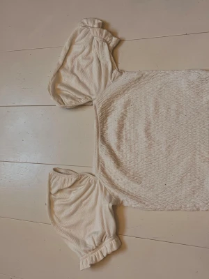 Vit topp med puffärmar och struktur - Söt vit topp med puffiga korta ärmar och lätt struktur i tyget. Fyrkantig ringning och resår vid ärmslut ger en trendig och feminin look. Perfekt att styla med jeans eller kjol för en fräsch outfit.