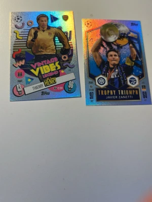 Glittriga fotbollssamlarbilder Topps - Två glittriga fotbollssamlarbilder från Topps, en med motiv av Thierry Henry och texten 'Vintage Vibes Legend', och en med Javier Zanetti och texten 'Trophy Triumph'. Båda har färgstarka detaljer och holografisk yta.