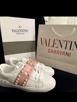 Valentino Garavani vita sneakers - Säljer ett par vita sneakers från Valentino Garavani med ljusrosa band och ikoniska nitar i metall. Skorna har en chunky sula med mönster och detaljerade nitar även baktill. Tillverkade i slätt läder och har en modern, stilren look.