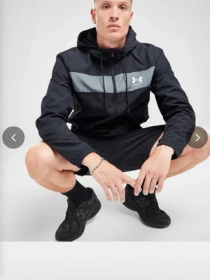Svart träningsjacka från Under Armour - Snygg svart träningsjacka från Under Armour med grått band över bröstet och vit logga. Jackan har huva, dragkedja och är tillverkad i lätt material som passar perfekt för träning eller sportiga aktiviteter. Knappt använd pris kan diskuteras 