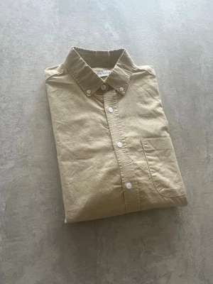 Beige skjorta från Studio Total - Klassisk beige skjorta från Studio Total med button-down krage och vita knappar. Skjortan har en bröstficka och är långärmad. Tillverkad i mjuk bomull för en clean och stilren look. Använd någon enstaka gång. Hör av er vid frågor!
