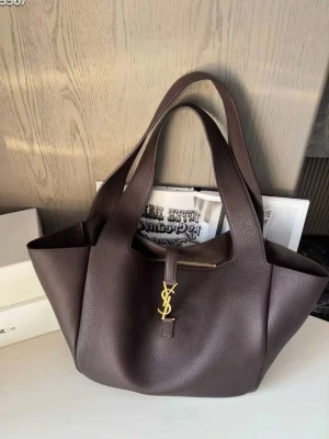 Mörkbrun YSL handväska i läder - Snygg mörkbrun handväska från YSL i mjukt, strukturerat läder. Väskan har dubbla handtag och ett guldfärgat YSL-emblem framtill. Rymlig modell med stilren design som passar perfekt till alla dina outfits.