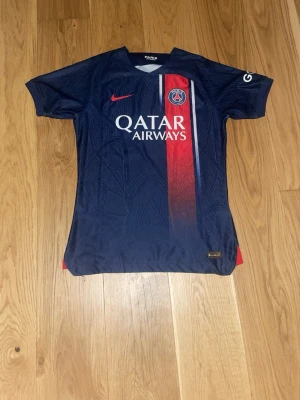 PSG matchtröja blå/röd Nike - Snygg PSG matchtröja i blått med röd vertikal rand och vita detaljer. Tröjan har klubbmärke, Nike-logga och Qatar Airways tryckt på bröstet. Tillverkad i lätt och ventilerande material, perfekt för fotboll eller träning.