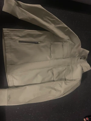 Beige overshirt från J.Lindeberg - Stilren mörk grön  overshirt från J.Lindeberg med två bröstfickor och en snedställd dragkedjeficka. Overshirten har en hög krage och diskret logga på ärmen. Tillverkad i ett slätt, tåligt material som ger en modern look.