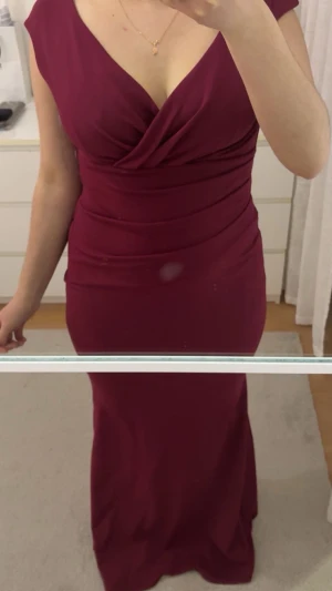 Vinröd balklänning från Goddiva - Elegant vinröd långklänning från Goddiva med omlott framtill och djup v-ringning. Klänningen är ärmlös och har figurnära passform med draperade detaljer i midjan. Perfekt för speciella tillfällen när du vill känna dig extra snygg. Har endast använt den 1 gång!!!! Pris kan diskuteras 