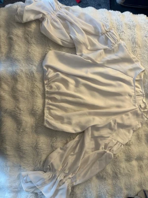 Vit satinblus med puffärmar - Stilren vit blus i glansigt satinmaterial med bred resår i midjan och voluminösa puffärmar. Ärmarna är långa och avslutas med resår och volangdetalj. Perfekt för dig som gillar trendiga och feminina plagg.