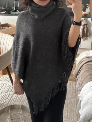 Mörkgrå ponchosjal med fransar - Säljer en mörkgrå ponchosjal med hög krage och långa fransar längs kanterna. Sjalen har en mjuk och följsam känsla, perfekt att slänga över axlarna för en chill och snygg look. Materialet känns som en skön stickad blandning.