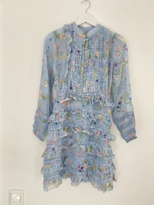 Blommig volangklänning i pastellblått - Supersöt kort klänning i pastellblått med blommigt mönster, volanger och spetsdetaljer. Långärmad modell med knappar framtill och hög krage. Perfekt för sommarens alla tillfällen!