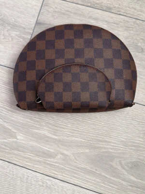 Brun sminkväska necessär med rutmönster - Snygg sminkväska i brunt och mörkbrunt rutmönster med röd insida. Väskan har en halvmåneform och dragkedja. Perfekt för att förvara smink eller småsaker. Endast använda som prydnad så i mkt bra skick. 