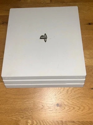 Vit PlayStation 4 Pro konsol - Säljer en vit PlayStation 4 Pro från Sony. Konsolen har en stilren, fyrkantig design med tre lager och PlayStation-loggan på ovansidan. Flera portar på baksidan för HDMI, USB och nätverk. Perfekt för gaming och streaming.