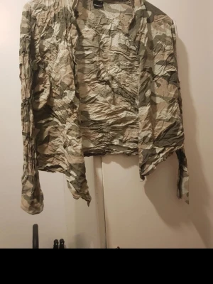 Camouflage vindjacka i beige och grön - Lätt och tunn vindjacka med camouflage-mönster i beige och gröna toner. Jackan har lång ärm och öppen front, perfekt för lager-på-lager. Tillverkad i syntetmaterial för en skön och smidig känsla. Storlek:40