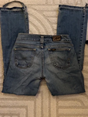 Blå jeans från Lee - Säljer ett par blåa jeans från Lee med klassisk femficksmodell och slitningar på benet. Jeansen har raka ben, låg midja och Lee-logga på bakfickan. Perfekta för en avslappnad och trendig look.