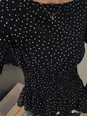 Svart prickig blus - Trendig svart blus med vita små prickar med resår i midjan. Blusen har långa ärmar och lätt volang nedtill. Jättefint skick 🤎
