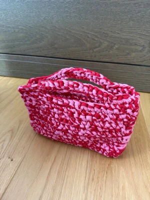 Handvirkad rosa och röd kuvertväska - Unik handvirkad kuvertväska i rosa och rött med inbyggt handtag. Väskan är gjord av tjockt garn som ger en cool och trendig look. Perfekt för dig som vill sticka ut med accessoarer i färg. 