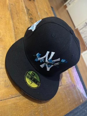 Svart New Era Yankees keps 59Fifty - Säljer en svart New Era 59Fifty keps med New York Yankees-logga framtill, dekorerad med blå blommor. På sidan finns ett broderat World Series-märke i blått och vitt. Klassisk flat brim och broderade detaljer. Perfekt för dig som vill sticka ut med snygga detaljer.