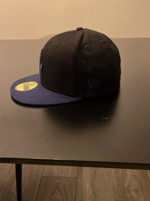 Svart LA-keps från New Era - Säljer en svart och mörkblå keps från New Era med LA-logga framtill. Modellen är 59FIFTY med platt skärm och broderade detaljer. Kepsen har officiella märken på sidan och klistermärke kvar på skärmen. Perfekt för dig som gillar streetwear och sportig stil.