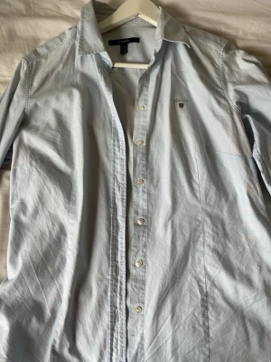 Ljusblå skjorta från Gant - Klassisk ljusblå skjorta från Gant med broderad logga på bröstet. Skjortan har knappar framtill, lång ärm och en tidlös design som passar till det mesta. Tillverkad i mjuk bomull för en skön känsla.