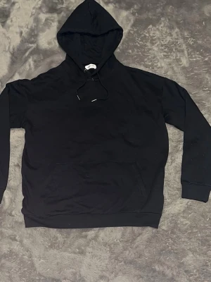 Svart basic hoodie med ficka - En svart hoodie med klassisk passform, stor huva och dragsko. Har en stor känguruficka framtill och ribbade muddar vid ärmar och midja. Perfekt för en avslappnad och stilren look.