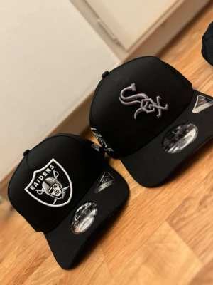 Svarta kepsar Raiders & White Sox - Två svarta snapback-kepsar från New Era, en med Raiders-logga i vitt och en med White Sox-logga i grått. Båda har böjd skärm och justerbar baksida. Klassisk streetwear-stil med broderade logotyper framtill. Perfekt för dig som gillar sportiga accessoarer. Både är helt helt nya