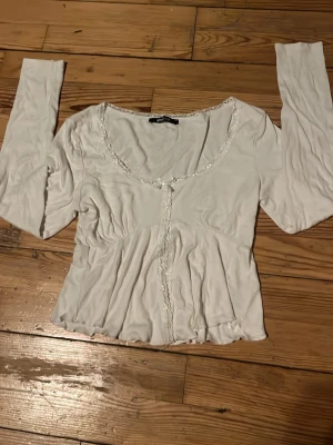 Vit långärmad blus med spetsdetaljer - Söt vit topp med långa ärmar och fina spetsdetaljer längs ringningen och framtill. Öppen/ har en slit vid mage. Modellen har en lätt volangulär midja och en något croppad passform 💗